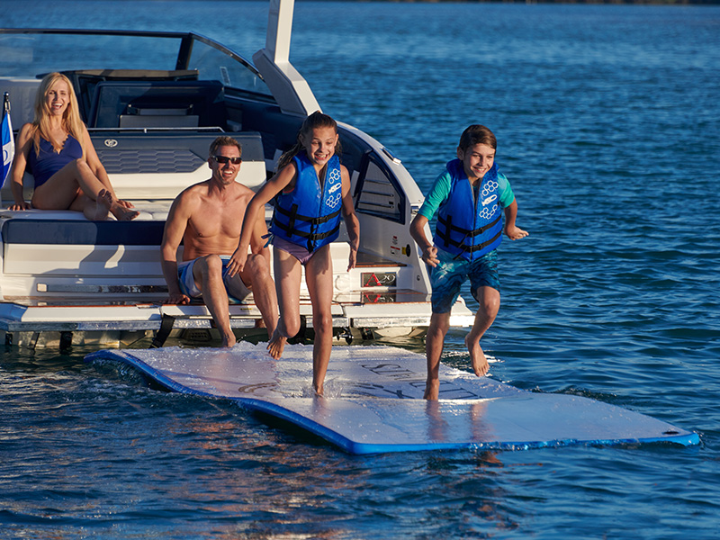 Two children running on the Cobalt A29 Splash and Stow inflatable float mat deployed from the stern into sun-drenched turquoise water