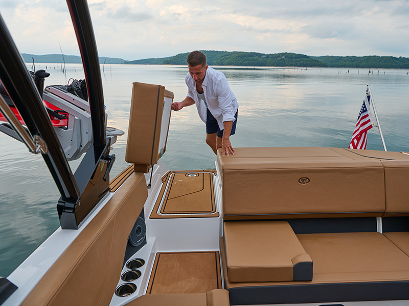 Man arranging the Cobalt CS23 optional walk-thru cushion in the aft seating area with tan upholstery on a calm evening lake