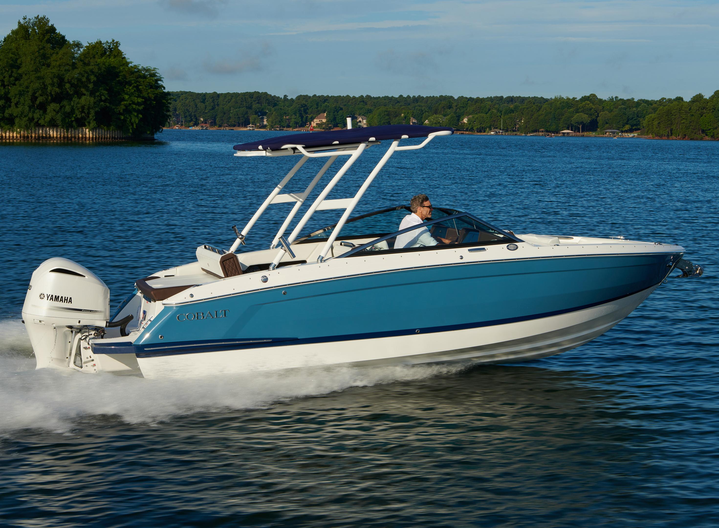 Person piloting a blue Cobalt outboard boat at speed on a calm freshwater lake surrounded by lush green trees