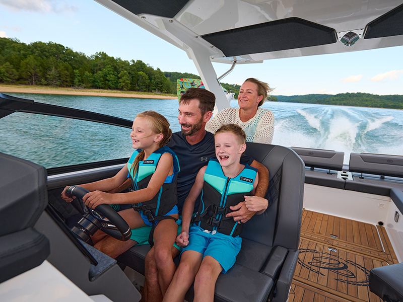 Family enjoying Cobalt R31 Sterndrive wide helm seat while underway on a scenic lake with life jacket-clad children at the wheel